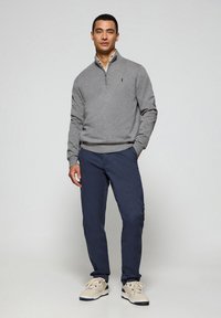 Sudadera gris con cremallera, pantalones azul marino y zapatillas beige. La sudadera tiene puños y dobladillo acanalados, y un pequeño emblema en el pecho. Texturas suaves visibles.