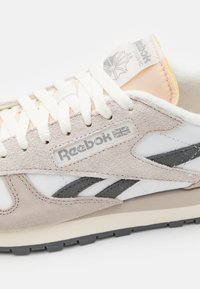 Zapatillas Reebok con un upper de gamuza gris y cuero blanco, cordones blancos y etiqueta de logo. La suela es beige con una banda de rodadura de goma negra.