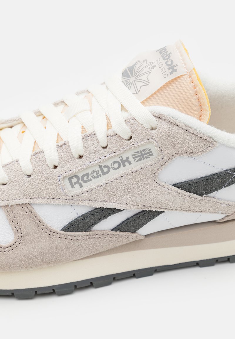 Baskets Reebok avec un dessus en daim gris et en cuir blanc, des lacets blancs et une étiquette de logo. La semelle est beige avec un profil en caoutchouc noir.