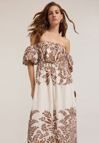 Robe maxi décolletée en tissu léger ; présente un fond blanc avec des motifs floraux marron intricats et une taille élastique.
