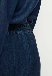 Tmavo modré denimové jogger nohavice majú elastický pás, jemnú textúru a zúžený strih. Tkanina vykazuje miernu variabilitu v tóne.