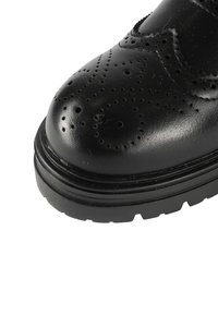 Scarpa di pelle nera con punta rotonda, perforazioni decorative tipo brogue e suola testurizzata. La scarpa ha una costruzione robusta e spessa.