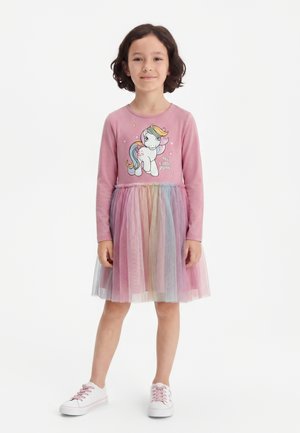 Pige iført en lyserød langærmet kjole med My Little Pony-grafik og en flerfarvet tylskørt, stående med hvide sneakers.