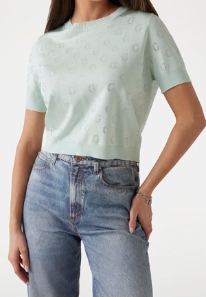 Kvinde iført en lysegrøn kortærmet striktrøje med et diskret "G"-mønster, kombineret med højtaljede, falmede blå jeans og et sølvarmbånd.