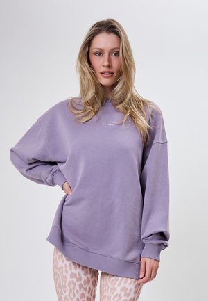 Lyshåret kvinde står med hånden på hoften iført en oversized lavendel sweatshirt med teksten "OH APRIL" og lyserosa bukser med leopardprint.