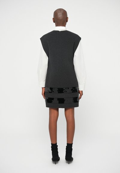 Vestido de lã cinza sem mangas com detalhes de lantejoulas pretas na orla, combinado com uma camisa branca. Botas pretas até aos tornozelos completam o visual.