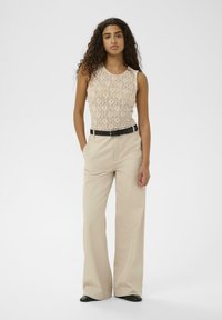 Młoda kobieta o kręconych włosach ubrana w beżowy, koronkowy top bez rękawów, szerokie spodnie z wysokim stanem w kolorze beżowym, czarny pasek i czarne buty, stojąca z rękami w kieszeniach.