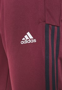 Pantalon Adidas marron avec des rayures verticales noires et le logo Adidas blanc sur la partie supérieure de la cuisse près d'une poche.