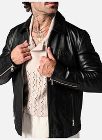 Veste en cuir noir au toucher lisse, avec des zips argentés et des accents côtelés, portée sur un haut en tricot beige avec un motif subtil.
