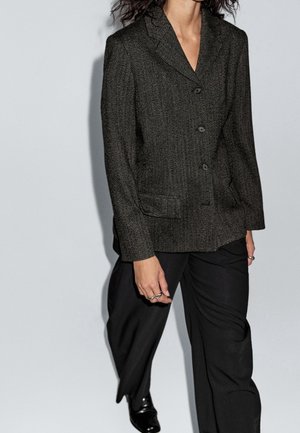 Personne portant un blazer texturé foncé avec des boutons, un pantalon noir à jambes larges et des chaussures noires marchant sur un fond clair.