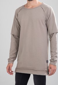 Sudadera de manga larga, color marrón claro, con dobladillo sin acabar, escote redondo y puños de canalé. Presenta un pequeño logo bordado en el pecho.