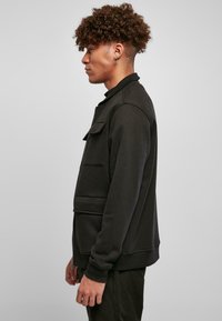 Črna zip-up sweatshirt s stojčkim ovratnikom, sprednjim žepom, rebrastimi manšetami in gladko teksturo. Obuta z ujemajočimi temnimi hlačami.