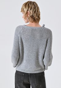 Maglione grigio lavorato a maglia con una vestibilità ampia, motivo testurizzato e un piccolo dettaglio con fascetta sul retro. Abbinato a jeans scuri.