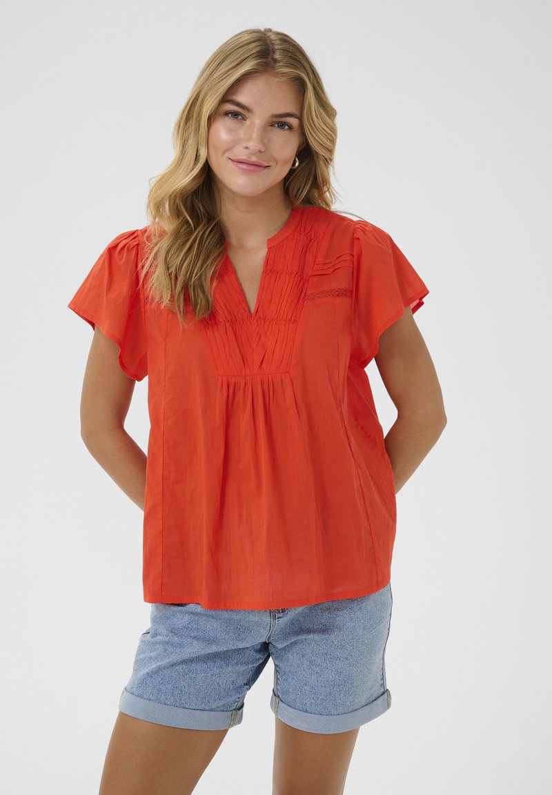 Blouse rouge à manches courtes avec un col en V, des détails plissés et une coupe décontractée. Associée à un short en denim bleu clair soigneusement retroussé aux poignets.
