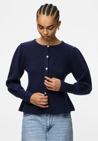 Jūras zilas adītas džemperis ar pūkainām piedurknēm, apaļu kakla izgriezumu un piecām dekoratīvām pogām. Auduma virsma ir teksturēta, ar ribotiem rakstiem.