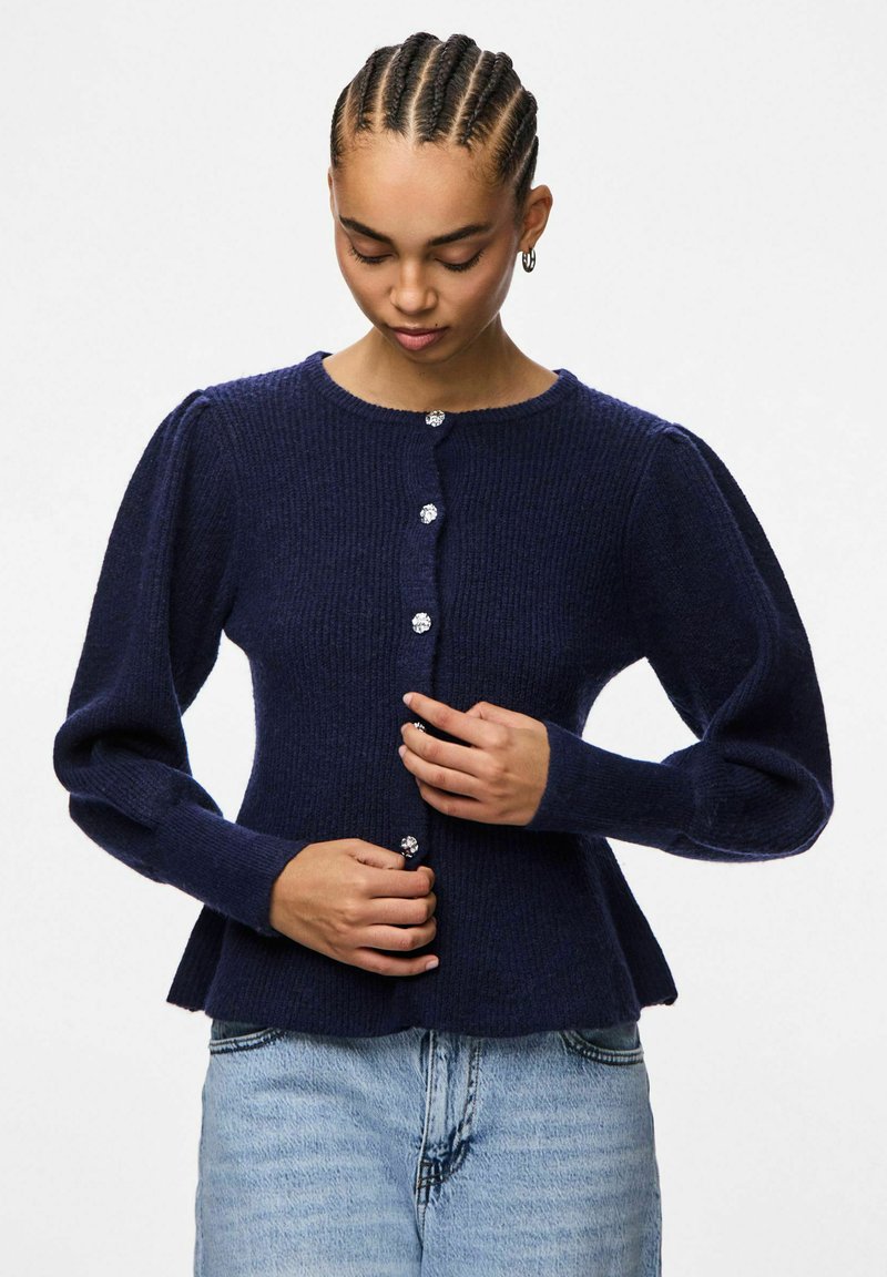 Jūras zilas adītas džemperis ar pūkainām piedurknēm, apaļu kakla izgriezumu un piecām dekoratīvām pogām. Auduma virsma ir teksturēta, ar ribotiem rakstiem.