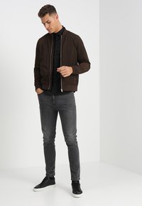 Braune Bomberjacke mit Reißverschluss, schwarzes Hemd darunter, graue Jeans und schwarze Sneakers. Glatter Stoff und lässiger Schnitt.
