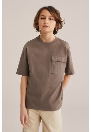 Jeune personne aux cheveux mi-longs portant un T-shirt marron ample avec une poche poitrine et un pantalon beige clair, debout devant un fond uni.