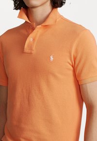 An orange polo shirt made of textured fabric, featuring a classic collar, two buttons, and a small white logo on the left chest.