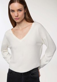 Maglione bianco a scollo a v con maniche lunghe, texture morbida, vestibilità rilassata, indossato su jeans neri. Design semplice senza pattern o abbellimenti visibili.