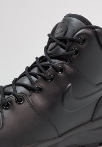 Kratki prikaz crnog visokog Nike čizme s kožnom teksturom, crnim vezicama provučenim kroz metalne nosače i utisnutim Nike swoosh logom.