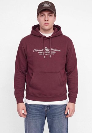 Un homme porte un sweat à capuche bordeaux orné d’un motif graphique, une casquette marron, un maillot de corps blanc et un jean bleu, sur fond blanc uni.