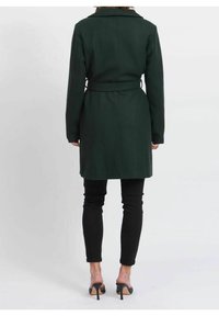 Dunkelgrüner Mantel mit tailliertem Gürtel, strukturiertem Kragen und langen Ärmeln. Glatte Textur, reicht bis zur Mitte des Oberschenkels, kombiniert mit schwarzer Hose.