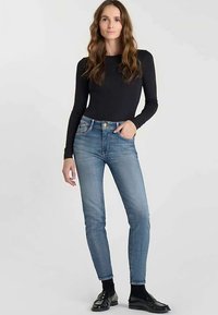 Haut noir à manches longues associé à un jean denim bleu clair ajusté. Le jean présente un design classique à cinq poches et une fermeture devant avec des boutons. Chaussures noires.
