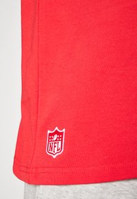 Rdeča bombažna majica s kratkimi rokavi, ki ima na robu bele NFL logotipne aplikacije in gladko, rahlo teksturirano površino.
