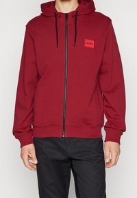 Červená zipová mikina vyrobená z měkké tkaniny, s kapucí, černými stahovacími šňůrkami a malou nášivkou loga na hrudi.