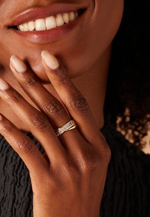 Bague en or avec un bandeau texturé, présentant une rangée de pierres claires incrustées. Une main avec des ongles naturels soigneusement manucurés repose contre le menton.
