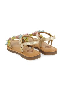 Sandalias planas doradas con adornos de cuentas multicolores, correas en el tobillo con hebillas y suelas marrones, mostradas desde un ángulo trasero.