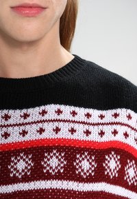Zbliżenie dolnej części twarzy i szyi osoby noszącej czarny, biały i czerwony sweter robiony na drutach z wzorami gwiazd i geometrycznymi.