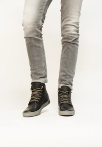 Zapatillas de cuero negras de caña alta con suelas de goma gris, con cordones redondeados y un acabado texturizado, combinadas con jeans slim-fit de color gris claro.