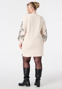 Beige jurk met zwarte bloemenborduursel op de mouwen, lange mouwen, zijsplitten, gecombineerd met zwarte kniehoge laarzen en zwarte panty’s.