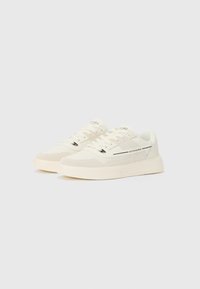 Cream sneakers with a textured upper, featuring perforated detailing, lace closure, and a chunky rubber sole. Black accent stripes included.