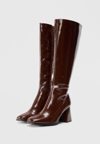 Botas castanhas de couro envernizado até ao joelho, com bico quadrado e salto robusto. Apresenta um fecho lateral com zipper e uma textura suave e brilhante.