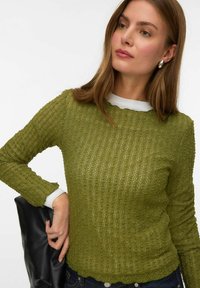 Maslinasto zelena majica dugih rukava s teksturiranim materijalom, rebrastim uzorkom i bijelim unutarnjim slojem na izrezu. Model drži crnu torbu.
