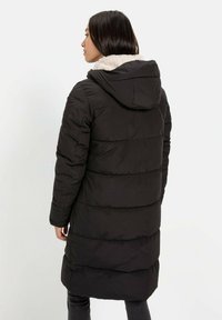 Zwarte gewatteerde jas met een capuchon, voorzien van een pluche kraag, horizontale stiksels en lange mouwen. Het ontwerp is middellang met een relaxte pasvorm.