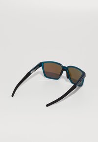 Gafas de sol azules y negras con montura rectangular, lentes marrones, patillas de goma y un logotipo en el lateral.