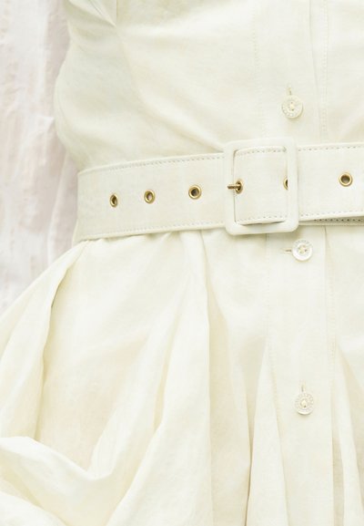 Κοντινό πλάνο μιας λευκής ζώνης με χρυσά κρικάκια που στερεώνεται γύρω από ένα πλισέ κρεμ φόρεμα με κουμπιά.