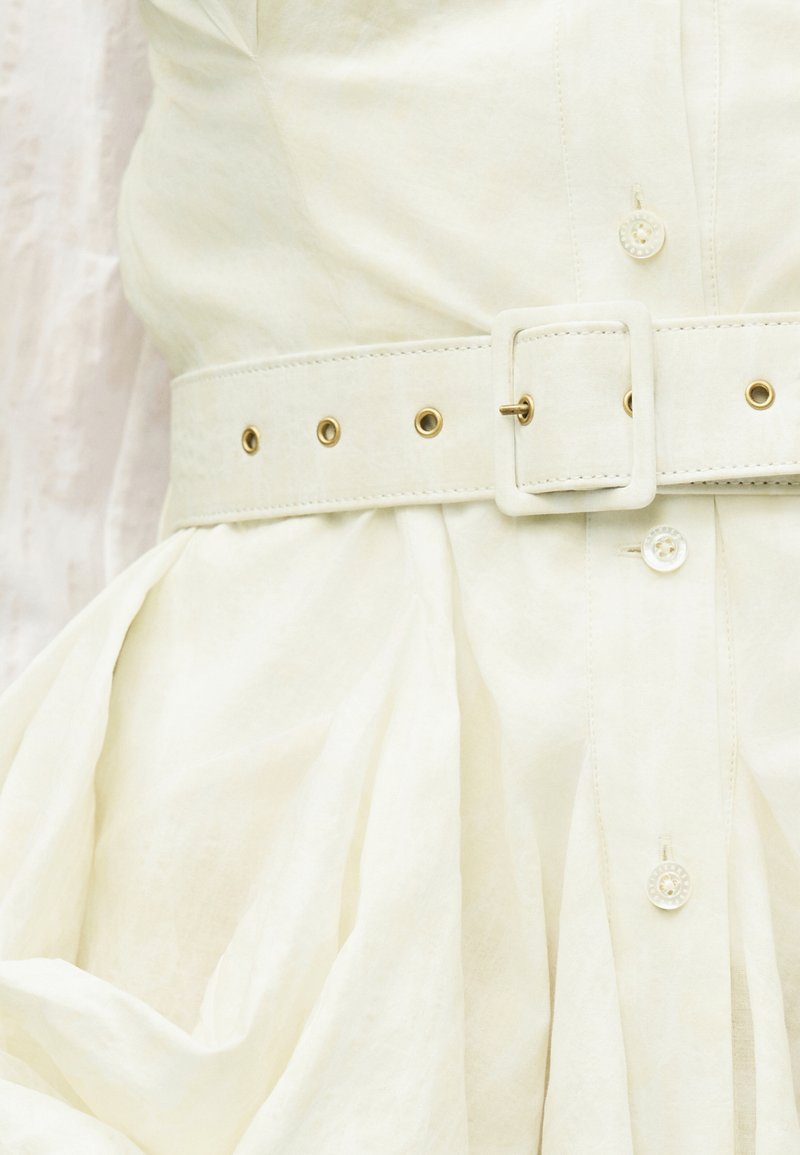 Gros plan d'une ceinture blanche avec œillets dorés attachée autour d'une robe plissée en tissu crème avec des boutons.