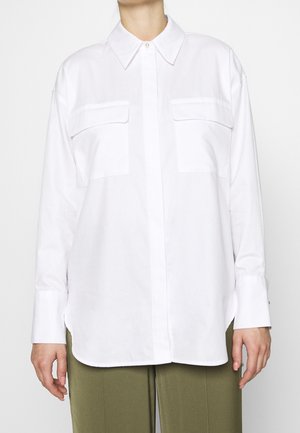 Chemise blanche à manches longues avec deux poches poitrine portée sur un pantalon vert olive, montrée sur une personne du cou aux hanches.