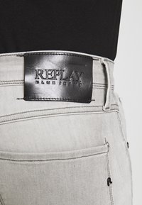 Šedé džínsy mají černou koženou nášivku s embosovaným nápisem "REPLAY BLUE JEANS". Jsou viditelné jemné švy a detaily kapes.
