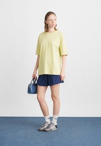 Mladá žena stojící na modrém koberci, oblečená v žlutém oversized tričku, námořnické plisované sukni, bílých ponožkách, stříbrných teniskách a držící modrou kabelku.