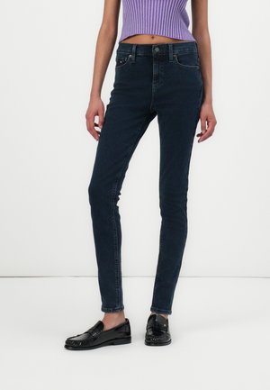 Vrouw die donkergroene skinny jeans draagt, een kort paars geribd mouwloos topje en zwarte leren loafers, staand tegen een witte achtergrond.