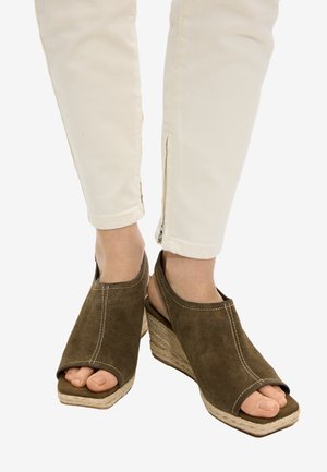 Stopy w brązowych zamszowych sandałach na koturnie z odkrytymi palcami i paskami na kostce, połączone z białymi spodniami z zamkami przy kostkach.