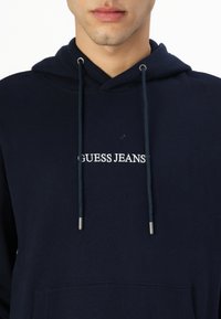 Sötétkék pamut kapucnis pulóver, elülső zsebbel, húzózsinórral ellátott kapucnival és fehér hímzett "GUESS JEANS" logóval a mellrészen.