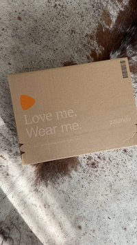Brown cardboard box with printed text “Love me. Wear me.” and orange logo. Located on a textured surface with brown and white patterns.