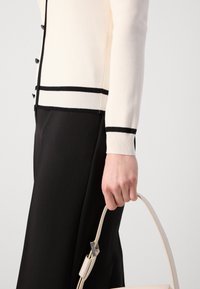 Cardigan crème à côtes avec des détails de bord noir, assorti à un pantalon noir. Une main tient un sac à main beige avec une fine bandoulière.